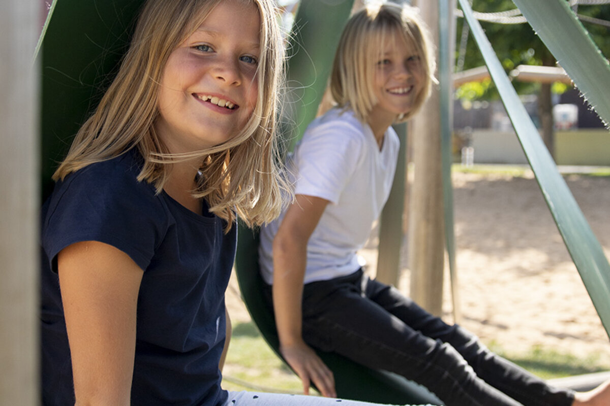 Schulhofplanung – Zwei lachende Mädchen sitzen in eibe Spielgeräten auf ihrem Schulhof.