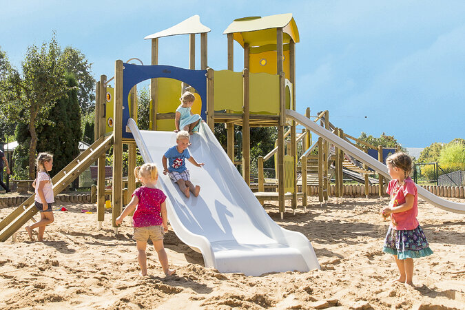 Kindergarten-Spielgeräte von eibe – bunte Spielanlage mit spielenden Kindern auf einer großen Sandfläche. 